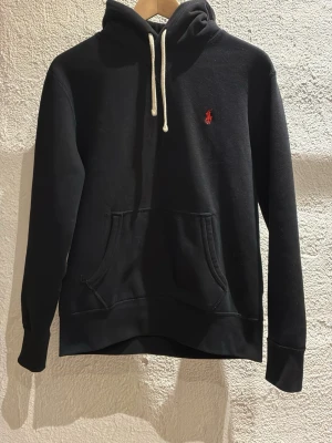 Svart hoodie från Polo Ralph Lauren - Svart hoodie från Polo Ralph Lauren med vit dragsko i huvan och röd broderad logga på bröstet. Klassisk känguruficka framtill och ribbade muddar vid ärmar och nederkant. Perfekt för en avslappnad och stilren look. Använd ett fåtal gånger ser ut som ny. Tveka inte på att höra av er vi funderingar. Pris går att diskutera 😄