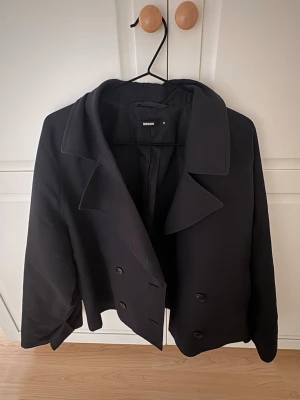 Svart jacka från Bikbok - Stilren svart rock från Bikbok i kortare modell med dubbelknäppning och bred krage. Klassisk design som passar perfekt till lager-på-lager och har en snygg, rak siluett. Tillverkad i ett mjukt tyg med slät yta och helfodrad insida. Köpt för 599kr.