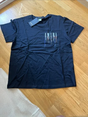 Svart AMIRI t-shirt med tryck - Svart t-shirt från AMIRI med färgstarkt tryck av märkesnamnet och droppande färgeffekt både fram och bak. Klassisk passform och rund halsringning. Tillverkad i mjuk bomull, perfekt för en trendig och avslappnad stil.