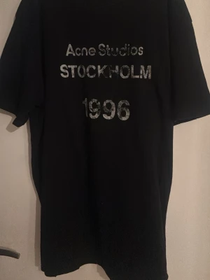 Svart Acne Studios t-shirt Stockholm - Cool svart t-shirt från Acne Studios med tryck i vitt och grått framtill där det står 'Acne Studios STOCKHOLM 1996'. Oversized passform och klassisk rund hals. Perfekt för dig som gillar minimalistisk och stilren design.
