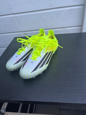 Adidas X Speedportal fotbollsskor - Säljer ett par Adidas X Speedportal fotbollsskor i vitt och neon-gult med svarta detaljer. Skorna har snörning, låg profil och är tillverkade i syntetmaterial för lätt vikt och bra grepp på planen. Perfekta för dig som vill ha snabbhet och stil på fotbollsplanen.
