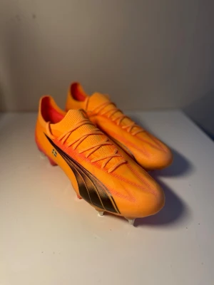 Puma orange Ultimate ultra SG - Snygga orangea fotbollsskor från Puma med svarta detaljer och svensk flagga på sidan. Skorna har metall-dobbar för bra grepp på planen och texten 'FOREVER FASTER' på hälen. Ovandelen är i syntetmaterial och designen är slimmad för maxad speed.