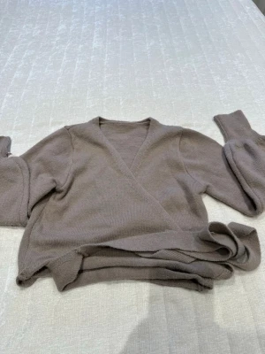 Beige omlottstickad tröja - Mysig beige stickad omlott-tröja med långa ärmar och knytband i midjan. Tröjan har en djup V-ringning och är tillverkad i mjukt material som ger en avslappnad och trendig look. Perfekt att styla över ett linne eller t-shirt.