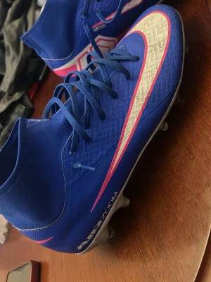 Blå Nike Mercurial fotbollsskor - Säljer ett par blå Nike Mercurial fotbollsskor med hög krage och snörning. Skorna har en stor vit och rosa Swoosh-logga på sidan, samt detaljer i rosa och vitt. Tillverkade i syntetmaterial med textur för bättre bollkontroll och dobbar för spel på gräs.