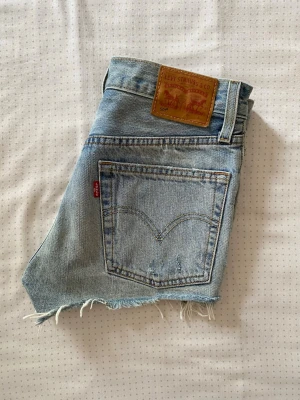 Äkta Levi's ljusblå jeansshorts  - Säljer ett par klassiska ljusblå jeansshorts från Levi's med råa, fransiga benslut och ikonisk läderpatch bak. Shortsen har fem fickor och den typiska röda Levi's-taggen på bakfickan. Perfekta för sommardagar och enkel att matcha. Priset kan diskuteras. Plagget säljs också på en annan plattform.