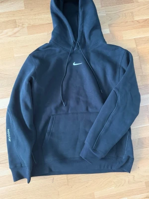 Svart Nike NOCTA hoodie - Säljer en svart hoodie från Nike NOCTA med huva och dragsnören. Framtill finns en liten vit Nike Swoosh och en stor magficka. På ärmen står det NOCTA och på ryggen finns diskreta broderade detaljer. Hoodien är i mjukt bomullsmaterial och har en avslappnad passform.
