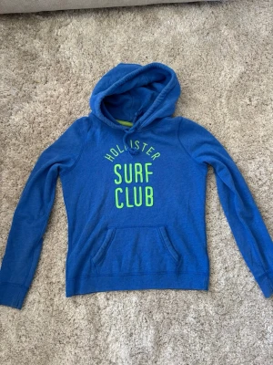 Blå Hollister hoodie med tryck - Snygg blå hoodie från Hollister med limegrönt tryck 'Surf Club' på bröstet. Klassisk känguruficka framtill och dragsko i huvan. Mjuk insida och ribbade muddar vid ärmslut och nederkant. Perfekt för en avslappnad och sportig stil.