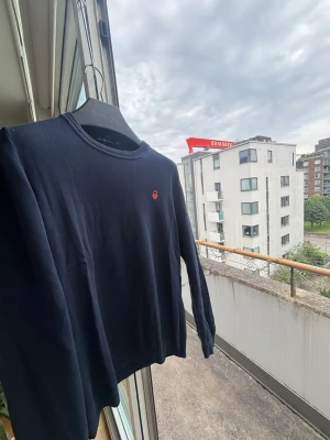 Mörkblå tröja från Sail Racing - Snygg mörkblå långärmad tröja från Sail Racing med rund halsringning och diskret orange logga på bröstet. Tröjan har ribbade muddar vid ärmslut och nederkant. Pris kan diskuteras!