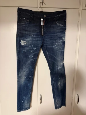 Mörkblå distressed jeans Dsquared2 - Säljer ett par mörkblå jeans från Dsquared2 med slitningar, färgstänk och en unik dragkedjedetalj fram. Jeansen har smal passform och klassisk femficksdesign. Perfekta för dig som gillar en edgy och trendig stil. Slim fit 