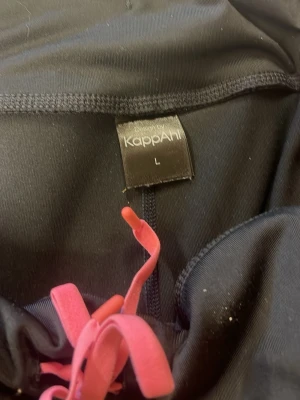 Svarta leggings från KappAhl L - Snygga svarta leggings från KappAhl i storlek L. De har coola rosa och röda detaljer med batikmönster längst ner på benen och matchande rosa snörning i midjan. Tillverkade i stretchigt material som sitter skönt och följer kroppen.