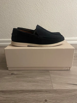 Selected Loafers - Snygga mörkblå loafers i mocka med klassisk design och kontrasterande vit sula. Skorna har rundad tå och diskreta sömmar längs ovandelen. Perfekta för en stilren och avslappnad look. Passar till jeans eller chinos. Märkena går att få bort enkelt både från under sulan och på sidan av skorna med en borste.