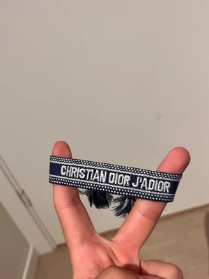 Christian Dior J'Adior armband - Snyggt marinblått tygarmband från Christian Dior med vit broderad text 'J'ADIOR'. Armbandet har dekorativa tofsar i ändarna och ett klassiskt, trendigt utseende som passar perfekt till din outfit.