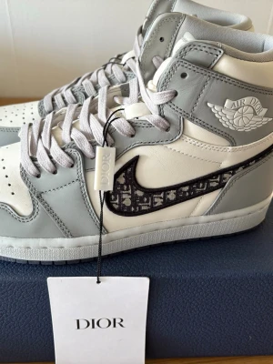 Air Dior x Jordan 1 High sneakers - Exklusiva Air Dior x Jordan 1 High sneakers i vitt och ljusgrått läder med ikoniska Dior-mönstrade Swoosh-loggor. Skorna har högt skaft, vita och grå skosnören samt Air Dior-detaljer på sidan. Perfekt för dig som vill sticka ut med lyxig streetwear.