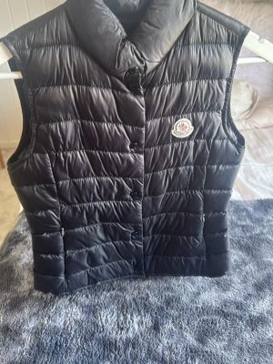 Svart dunväst från Moncler - Snygg svart dunväst från Moncler med klassisk quiltad design och hög krage. Västen har tryckknappar framtill och Monclers ikoniska logotyp på bröstet. Perfekt för lager-på-lager och håller dig varm med lättviktig dunfyllning.