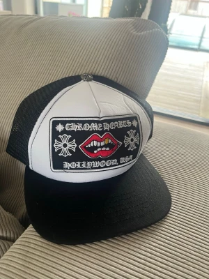 Chrome Hearts keps - Snygg truckerkeps från Chrome Hearts i svart och vitt med broderad patch framtill med text, blommor och röda läppar. Baksidan har justerbar snapback och meshmaterial för extra ventilation. Märkeslogga broderad även bak.