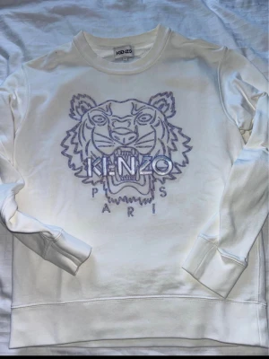Vit Kenzo sweatshirt med tigertryck - Vit sweatshirt från Kenzo med ikoniskt broderat tigerhuvud och texten 'KENZO PARIS' framtill. Tröjan har rund halsringning, långa ärmar och ribbade muddar. Perfekt för dig som gillar streetwear och vill sticka ut med en klassisk design.
