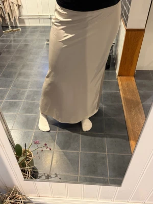 Beige långkjol i satin material - Säljer en stilren beige långkjol med rak passform. Kjolen har en lite skrapa där fram, men är inget synligt när man har den på sig som ni ser! följsamt material som ger en clean look. Perfekt att matcha med både sneakers och boots.