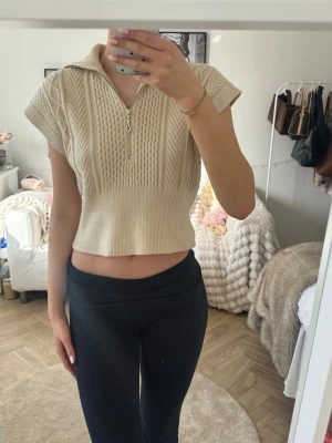 Beige stickad crop top med dragkedja - Trendig beige stickad crop top med korta ärmar och halv dragkedja framtill. Snygg kabelstickad design och bred krage som ger en cool look. Perfekt att styla med höga jeans eller kjol. Mjuk och skön kvalitet.