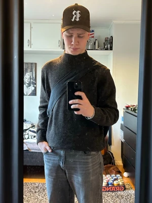 Mörkgrå stickad tröja med ribbad krage - Säljer en mörkgrå stickad tröja med bred ribbad krage och ribbade muddar vid ärmslut och nederkant. Tröjan har en klassisk och enkel design, perfekt för lager-på-lager och chill stil. Materialet ser ut att vara ull eller ullblandning.