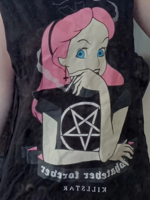 Svart t-shirt Killstar Alice tryck - Svart t-shirt från Killstar med stort tryck av en tecknad Alice med rosa hår, blå ögon och pentagram på bröstet och Killstar-logga nedtill. T-shirten är ärmlös och har edgy goth-vibe.