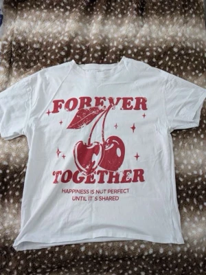 Vit t-shirt med körsbärstryck - Vit t-shirt med rött tryck framtill som föreställer körsbär och texten 'Forever Together'. T-shirten har korta ärmar och en klassisk passform. Perfekt för dig som gillar grafiska prints och en avslappnad stil.