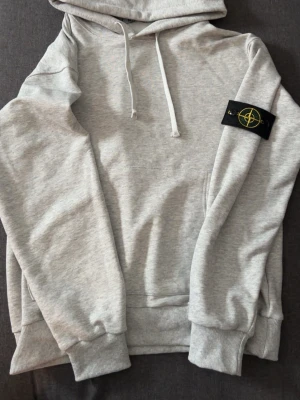 Grå hoodie från Stone Island - Snygg ljusgrå hoodie från Stone Island med klassisk patch på ärmen. Hoodien har huva med dragsko, magficka och ribbade muddar vid ärmslut och midja. Perfekt för en avslappnad streetwear-look.