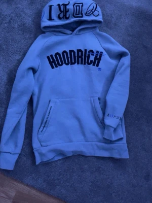 Ljusgrå hoodie från Hoodrich - Pris kan diskuteras) Snygg ljusgrå hoodie från Hoodrich med stor svart logga på bröstet och coola tryck på huvan och ärmen. Klassisk känguruficka framtill och mjukt material som känns skönt mot huden. Perfekt för en avslappnad streetwear-look.