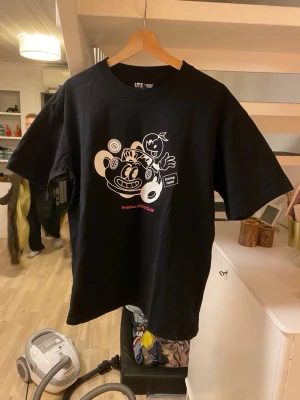 Svart t-shirt med grafiskt tryck UTI - Cool svart t-shirt från UTI med ett stort grafiskt tryck på framsidan i vitt och rött. Trycket har en lekfull design med figurer och text. T-shirten har rund hals och korta ärmar, perfekt för dig som gillar streetstyle och unika prints.