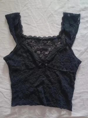 Svart spets topp med rosett💝 - Snygg svart topp i spets med v-ringning och liten rosett framtill. Toppen har breda axelband och är genombruten med blommigt spetsmönster. Perfekt för dig som gillar en feminin och trendig stil. Är lite trasig nere av toppen ---> sista bilden. Står ingen storlek men skulle säga att den är xs/s. 