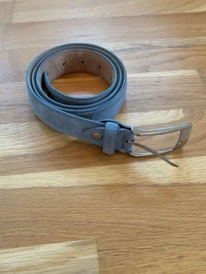 Grått mockabälte med silverfärgat spänne - Säljer ett stilrent grått bälte i mocka med ett klassiskt silverfärgat metallspänne. Bältet har en enkel design och flera hål för justering. Perfekt för att ge din outfit en snygg touch. Säljer för 219kr men är öppen för prisförslag. Bältet är 105cm och i väldigt bra skick.