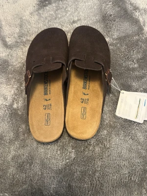 Bruna Birkenstock Boston tofflor - Mörkbruna Birkenstock Boston tofflor i mocka med klassisk slip-in design och justerbart spänne i metall. Fotbädden är i kork och mocka för extra komfort. Perfekta för en chill och avslappnad stil.