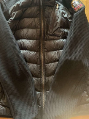 Svart pufferjacka från Parajumpers - Snygg svart pufferjacka från Parajumpers med quiltad front och släta ärmar i mesh-liknande material. Jackan har dragkedja framtill, bröstficka med dragkedja och klassisk Parajumpers-logga på axeln. Perfekt för kalla dagar.