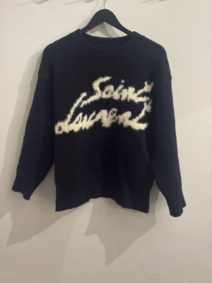 Saint Laurent stickad tröja - Cool svart stickad tröja från Saint Laurent med vit fuzzy logotyp framtill. Tröjan har rund halsringning, långa ärmar och ribbade muddar vid ärmslut och nederkant. Perfekt för dig som gillar statement-plagg och exklusiv streetstyle.