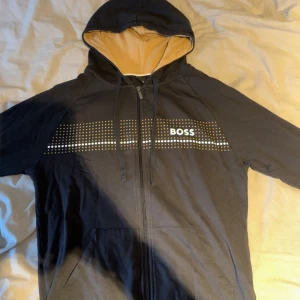 Svart hoodie från BOSS med dragkedja - Svart hoodie från BOSS med vitprickigt mönster och logga över bröstet. Tröjan har huva med snörning, dragkedja framtill och fickor. Insidan av huvan är beige för en snygg kontrast. Perfekt för en chill och stilren look.