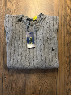 Grå kabelstickad tröja Polo Ralph Lauren - Snygg grå kabelstickad tröja från Polo Ralph Lauren med rund halsringning och klassisk svart broderad logga på bröstet. Tröjan har långa ärmar och ribbade muddar vid ärmslut och nederkant. Perfekt för lager på lager och stilren vardagslook.