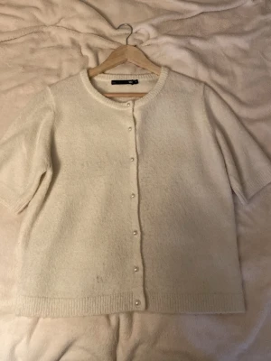 Vit kofta med pärlknappar från 157 - Söt vit kofta från 157 med korta ärmar och rund halsringning. Koftan har pärlknappar framtill och är stickad i ett mjukt material. Perfekt att slänga över en topp för en clean och enkel look.