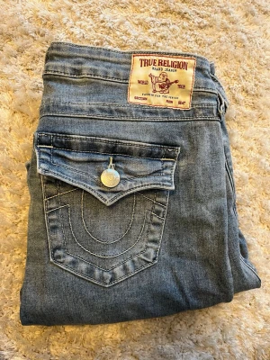 True Religion utsvängda jeans - Snygga blå jeans från True Religion med klassisk fickdesign och utsvängda ben. Jeansen har en ljus tvätt och detaljerade sömmar samt en ikonisk logga på bakfickan. Perfekta för dig som gillar en retro och avslappnad stil.