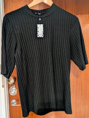 Svart ribbad topp med halvlång ärm - Stilren svart topp med ribbad struktur. Toppen har en högre rund halsringning och är tillverkad i ett mjukt, stretchigt material som ger en bekväm passform. 