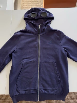  C.P. Company hoodie - Säljer en mörkblå hoodie från C.P. Company med dragkedja framtill och två stora fickor. Huvan har inbyggda glasögon, vilket ger en unik och cool look. Tillverkad i mjukt bomullsmaterial och har diskret logga på bröstet. Perfekt för dig som vill sticka ut.