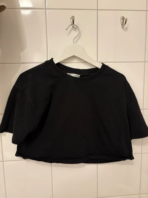Svart croppad t-shirt från Zara - Svart croppad t-shirt från Zara med rund halsringning och korta ärmar. Enkel och stilren design i mjuk bomull, perfekt till jeans eller kjol. Snygg basic som funkar året runt.