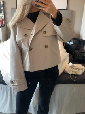 Kort beige trenchcoat med stora knappar - Säljer en helt ny snygg kort trenchcoat i beige med dubbelknäppning och stora bruna knappar. Jackan har bred krage och dekorativa knappar på ärmsluten. Perfekt för dig som vill ha en trendig och stilren look.