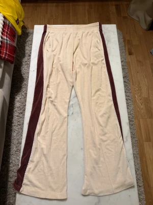 Beige mjukisbyxor med vinröda sidor jaded London - Jaded London. Säljer ett par beige mjukisbyxor med breda vinröda paneler längs sidorna. Byxorna har raka ben, resår i midjan och dragkedjeförsedda fickor. Materialet är mjukt velour och det finns en broderad logga på ena benet. Finns ett litet hål på ena benet längst ner. Kan visa bild 