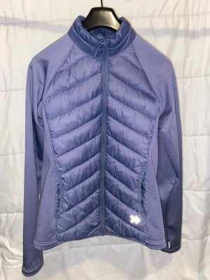 Lila pufferjacka från Columbia - Snygg lila pufferjacka från Columbia med quiltade partier framtill och baktill. Jackan har dragkedja hela vägen, hög krage och tumhål vid ärmsluten. Materialet är en mix av glansigt och stretchigt tyg för extra komfort och värme.