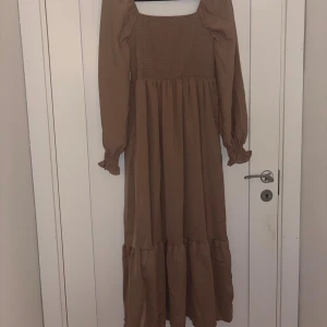 Brun långklänning med puffärmar - Snygg brun långklänning med smockad överdel och långa puffärmar. Klänningen har volangdetalj nertill och en fyrkantig ringning. Perfekt för dig som gillar en romantisk och trendig stil.