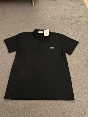 Svart pikétröja från Lacoste - Klassisk svart pikétröja från Lacoste med broderad krokodil-logga på bröstet. Tröjan har korta ärmar, krage och knappar framtill. Perfekt för en stilren och sportig look.