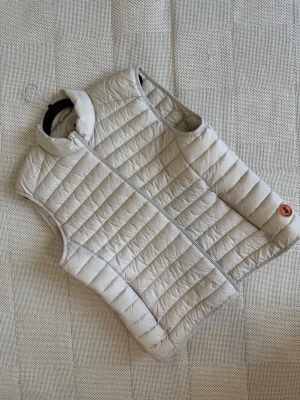 Ljusbeige dunväst med ståkrage - Snygg ljusbeige dunväst med quiltat mönster och ståkrage. Västen har dragkedja framtill och en diskret logotyp på sidan. Perfekt för lager-på-lager och enkel att matcha med olika outfits.