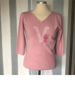 Rosa stickad tröja S - Snygg rosa stickad tröja  med V-logga och emblem framtill. Tröjan har v-ringning och trekvartsärmar samt ribbad nederkant och ärmslut. Perfekt för dig som gillar exklusiva och trendiga plagg med en feminin touch.