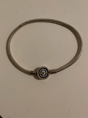 Silverfärgat armband med stenar - Snyggt armband i silverfärgad metall med en rund stendetalj i mitten, omgiven av gnistrande stenar. Armbandet har en stilren och modern design med ett flätat band och passar perfekt för dig som gillar eleganta accessoarer. En liten sten saknas men det märks knappt, annars i perfekt kondition.