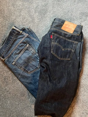Levi's jeans i två olika blå nyanser - Tja! jag säljer två par klassiska Levi's jeans i olika blå tvättar. Väldigt sällan använda. Ena paret är ett mörkblå 501s med kontrastsömmar och läderpatch bak där kan man även se storleken ena har en liten fläck som knappt syns vid bakfickan annars är de som nya, det andra paret är ljusare blått. Båda har raka ben med en 501 design och femficksdesign. Perfekta för en stilren look. 