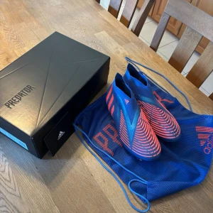 Adidas Predator blå/röda fotbollsskor - Säljer ett par Adidas Predator fotbollsskor i blått med röda detaljer och mönster. Skorna har en strumpliknande design utan snörning och levereras med en blå mesh-påse och originalkartong. Perfekta för dig som vill ha grym touch och kontroll på planen.